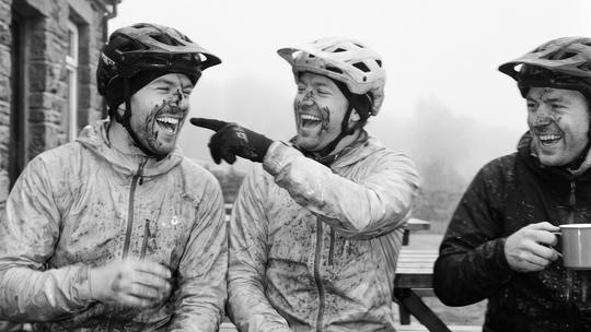 three muddy riders on a bench outside a café, one with a roll, all laughing