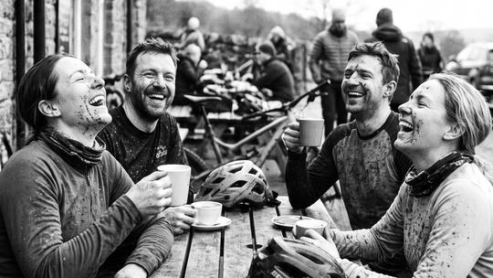 four muddy riders laughing over coffee outside a trail-centre café