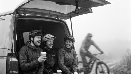 three riders with coffees on the back of a van as another rides past