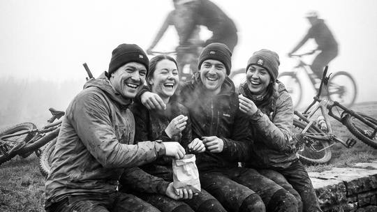 four riders sat on a stone wall sharing a bag of snacks, bikes in the grass behind